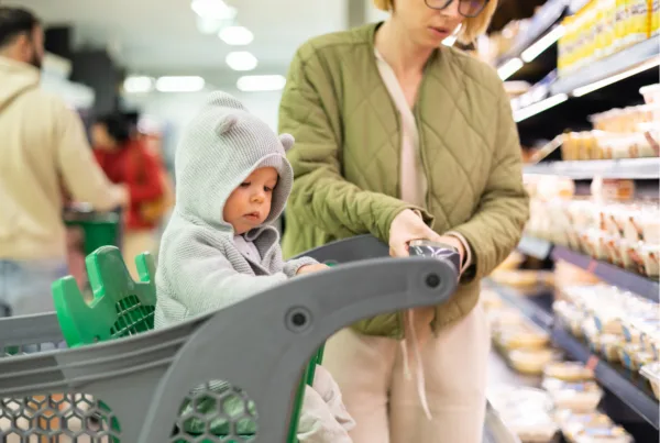 A mom and baby navigate the grocery store with their cart to purchase food items.