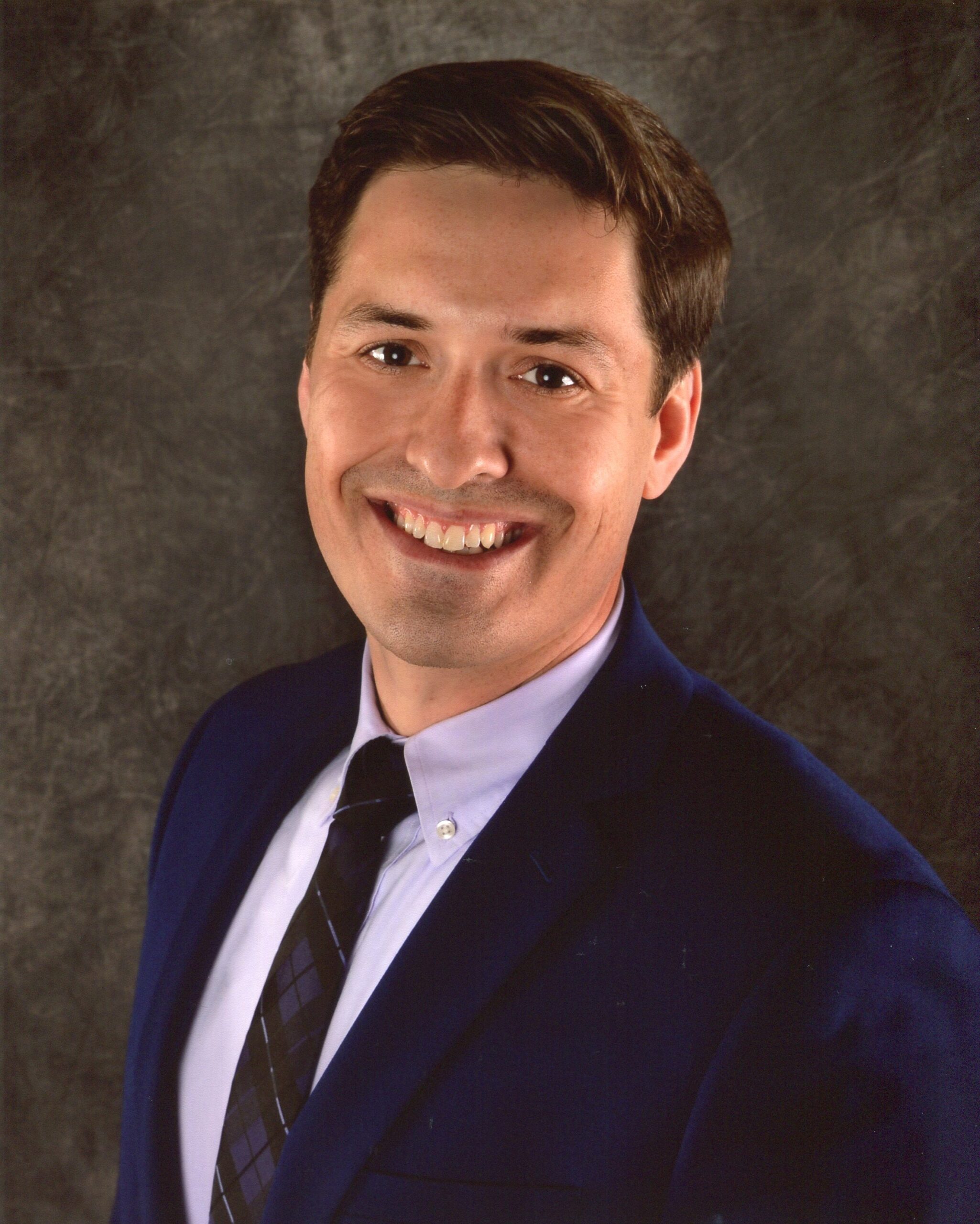 A smiling man in a black coat, white shirt, and black tie.