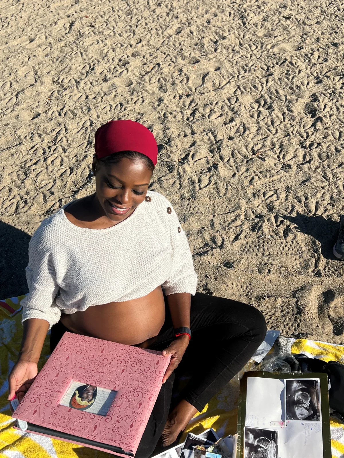 A pregnant woman in a white shirt and red hat sitting on a sandy beach