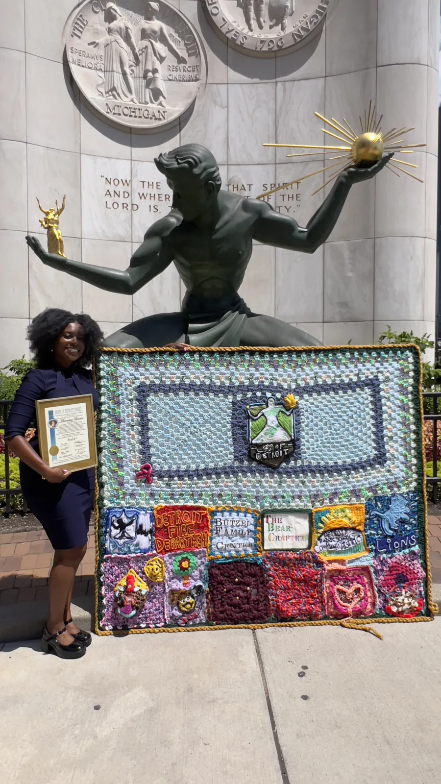 A woman holding a certificate and displaying a blanket she made in front of the Spirit of Detroit statue