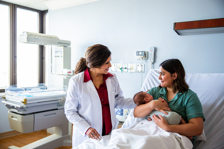 Pediatrician of Indian ethnicity smiling with a newborn baby and his mother in a hospital bed