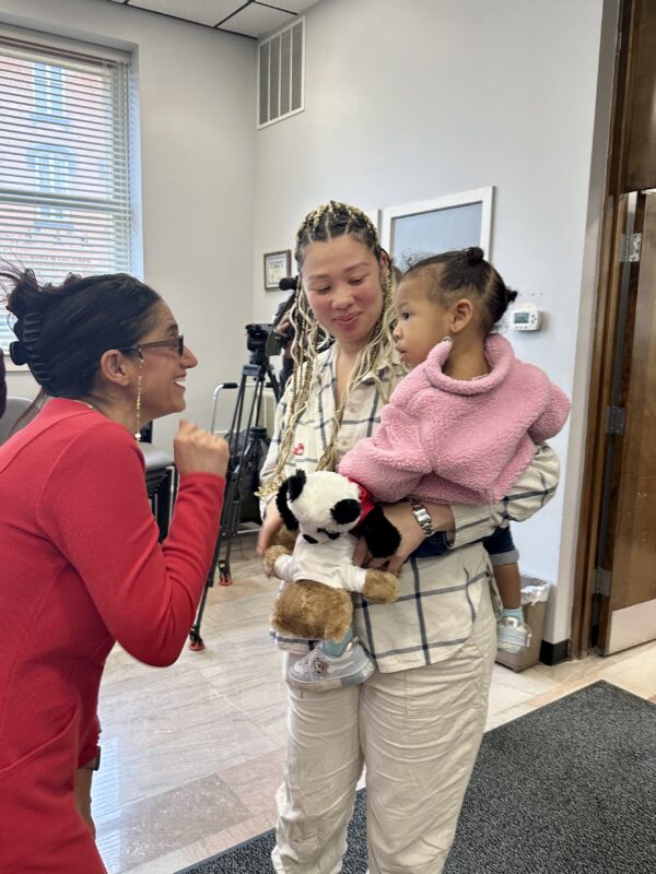 A mom holds a baby in a pink coat while a woman in a red dress smiles at her